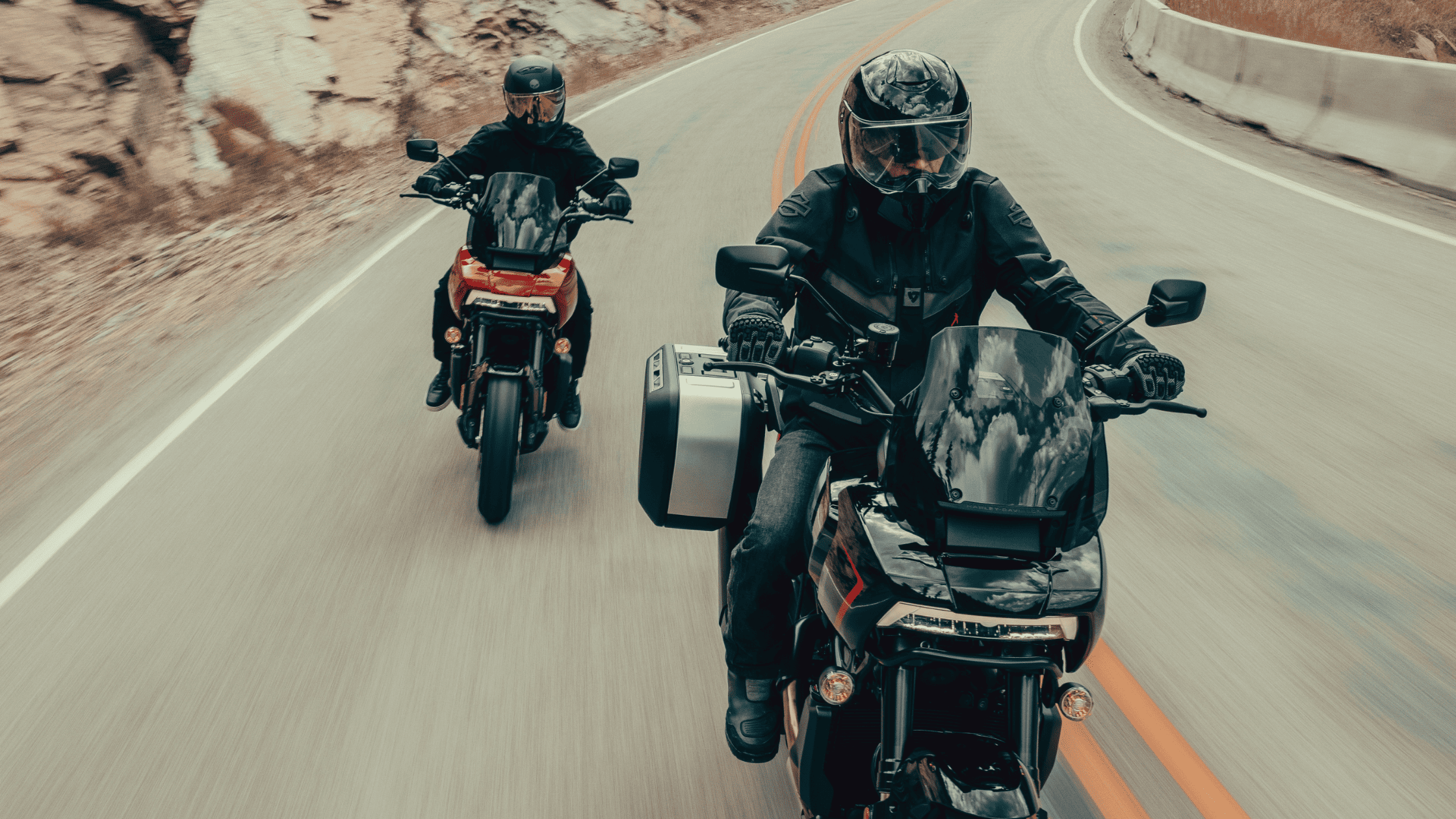Two motorcyclists in full riding gear navigate a winding mountain road, riding adventure touring motorcycles equipped with windshields and side panniers. The background features rocky terrain and an open road, emphasizing long-distance travel and adventure.