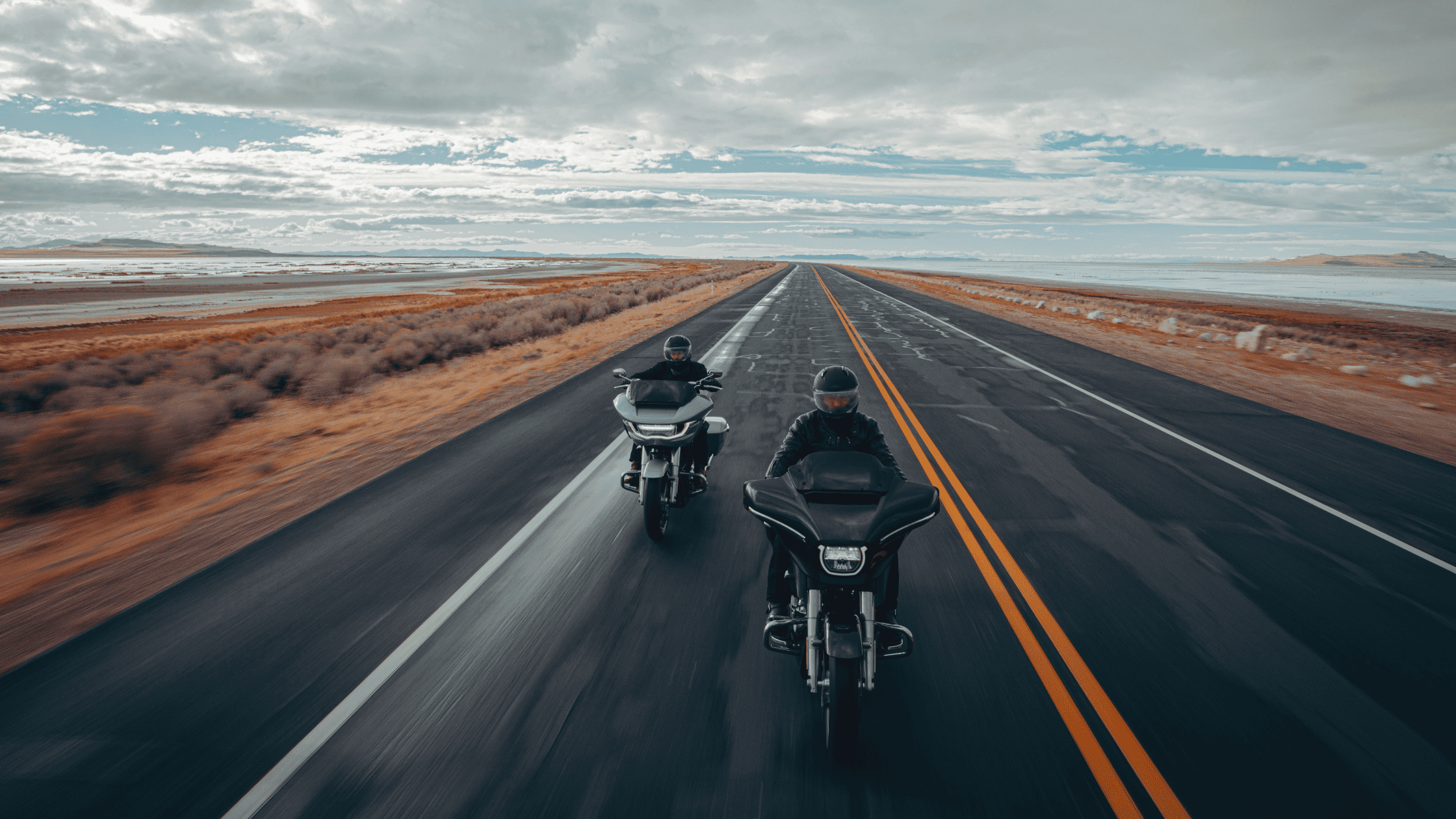 Two riders on sleek, fully-equipped Grand American Touring motorcycles travel down an expansive open highway under a partly cloudy sky. The road stretches endlessly ahead, symbolizing freedom, long-distance travel, and the essence of touring motorcycles.