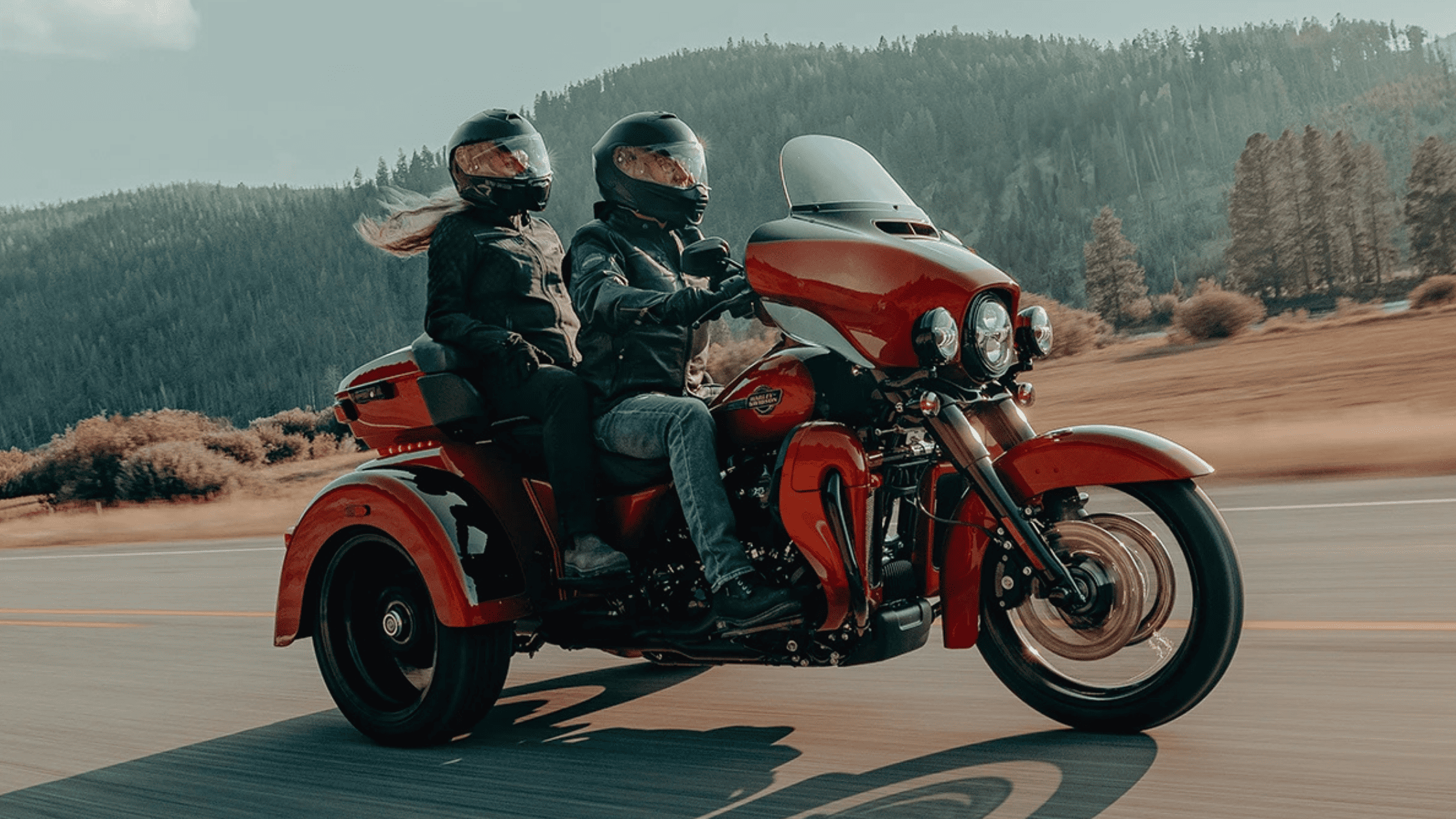 A rider and passenger cruise down a scenic highway on a bold red trike motorcycle, set against a backdrop of rolling hills and open countryside. The three-wheeled design offers stability and comfort, making it ideal for long rides and relaxed touring.
